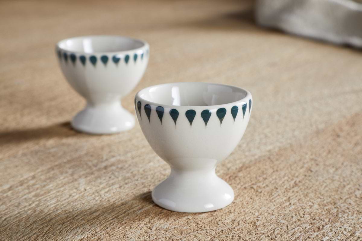 The nkuku Juniper Drop Egg Cups - Cream & Juniper (Set of 2) feature a dark blue teardrop pattern on white ceramic, ideal for adding style to your breakfast table.