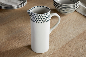 The nkuku Juniper Drop Jug – Cream & Juniper – Large, a white ceramic jug with a handle and blue teardrop accents near the rim, sits on a wooden table among stacked matching bowls and plates.