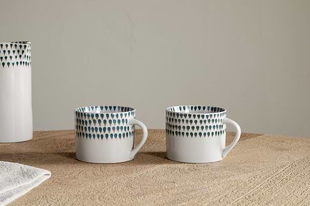 Two nkuku Juniper Drop Mugs in cream and juniper, featuring unique handles and signature drop design, rest side by side on a wooden surface. A matching vase and a white cloth are partially visible to the left. Set of 2 (Small).