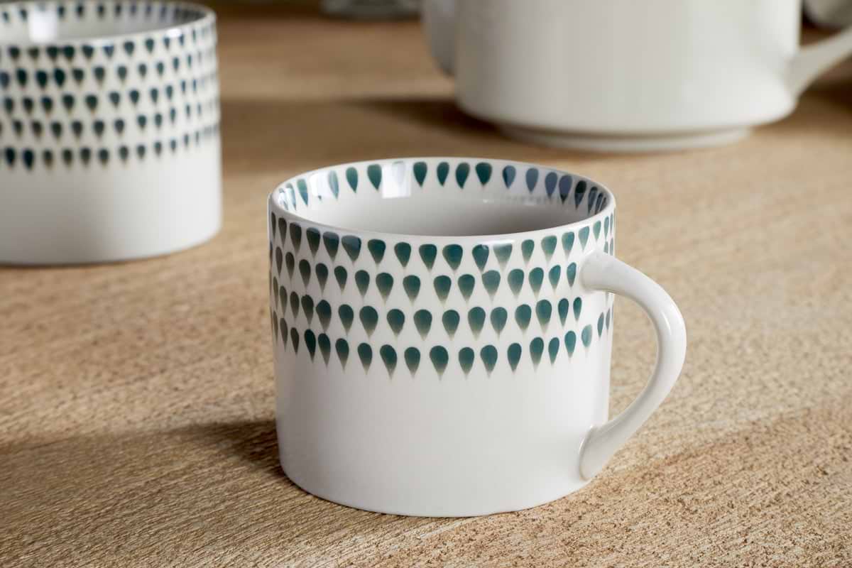 The nkuku Juniper Drop Mug - Cream & Juniper - Small (Set of 2) features a teardrop pattern in cream and juniper tones, displayed on a wooden surface with another matching mug and part of a teapot softly blurred in the background.