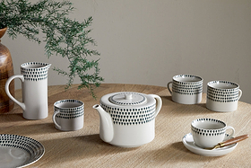 The nkuku Juniper Drop Mug - Cream & Juniper (Set of 2, small) features a white ceramic design with black dots and unique handles, sitting on a beige table. A green leafy plant in a vase decorates the background.