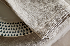 A close-up of the nkuku Junto Linen Napkin - Natural (Set of 4) draped over a table edge, partially covering a white plate with dark blue teardrop patterns.