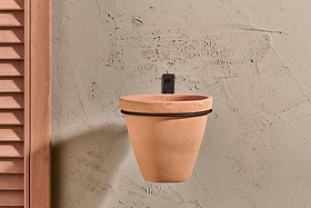 The Jara Single Wall Hung Planter by nkuku, a plain terracotta pot in a black metal holder, hangs on a textured beige wall. To the left is part of a wooden shutter, and the empty planter casts a faint shadow on the wall.