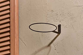 The nkuku Jara Single Wall Hung Planter is mounted on a textured beige wall, its metal ring casting an oval shadow. To the left, a partially visible brown wooden shutter with vertical slats adds contrast.