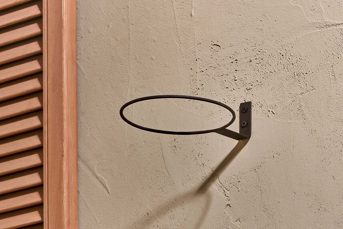 The nkuku Jara Single Wall Hung Planter is mounted on a textured beige wall, its metal ring casting an oval shadow. To the left, a partially visible brown wooden shutter with vertical slats adds contrast.