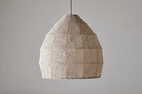 The nkuku Jahari Woven Jute Pendant - Natural hangs from the ceiling against a plain, light gray wall, offering rustic charm with its woven beige dome-shaped design.