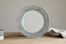 The nkuku Juniper Drop Dinnerware Set - Cream & Juniper (Set of 12) features white ceramic plates with dark blue teardrop patterns on the rim, shown here on a wooden table alongside stacked dishes, a folded beige cloth, and a matching cup.