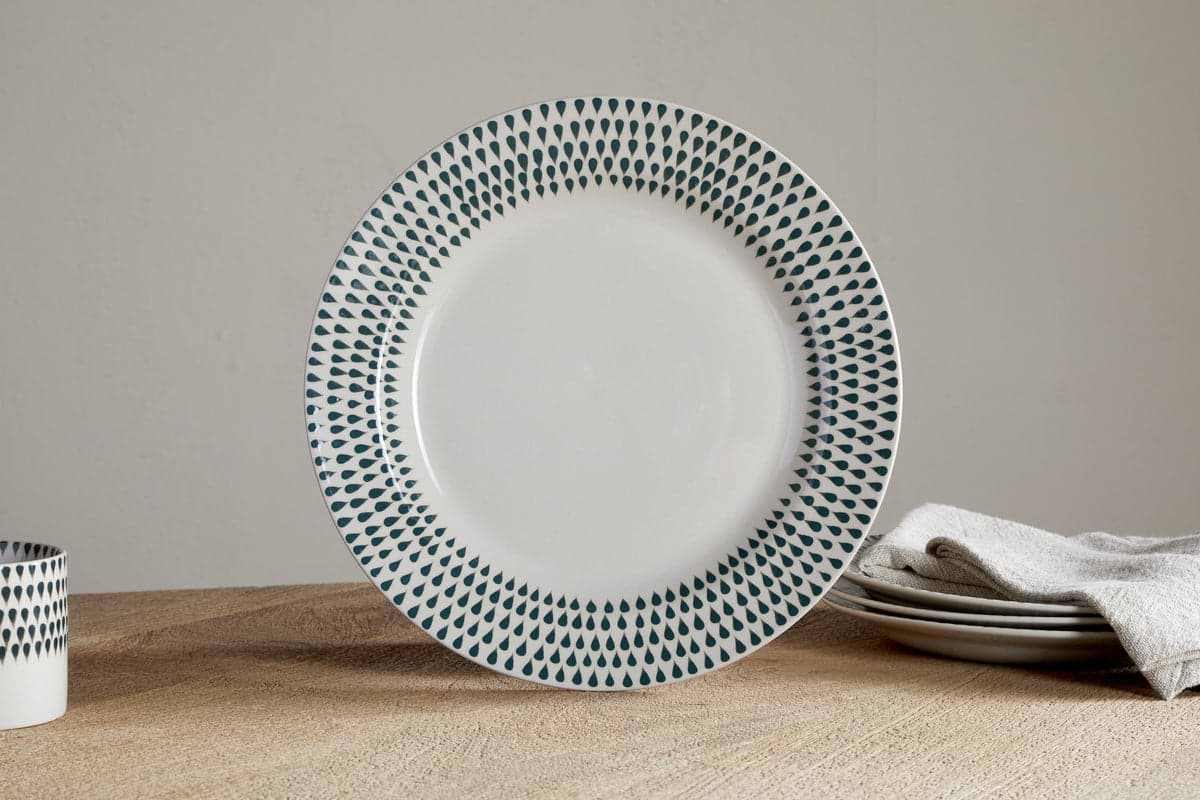 The nkuku Juniper Drop Dinner Plate in Cream & Juniper features a green drop pattern on white ceramic, shown upright beside stacked plates, a napkin, and a matching cup—ideal for modern table settings.