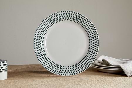 The nkuku Juniper Drop Dinner Plate in Cream & Juniper features a green drop pattern on white ceramic, shown upright beside stacked plates, a napkin, and a matching cup—ideal for modern table settings.