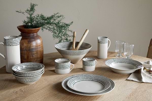 A set of nkuku Juniper Drop Dinner Plates in Cream & Juniper, featuring dark dotted patterns, is arranged on a wooden table with matching bowls, mugs, pitchers, glasses, and a brown vase with greenery in the background.