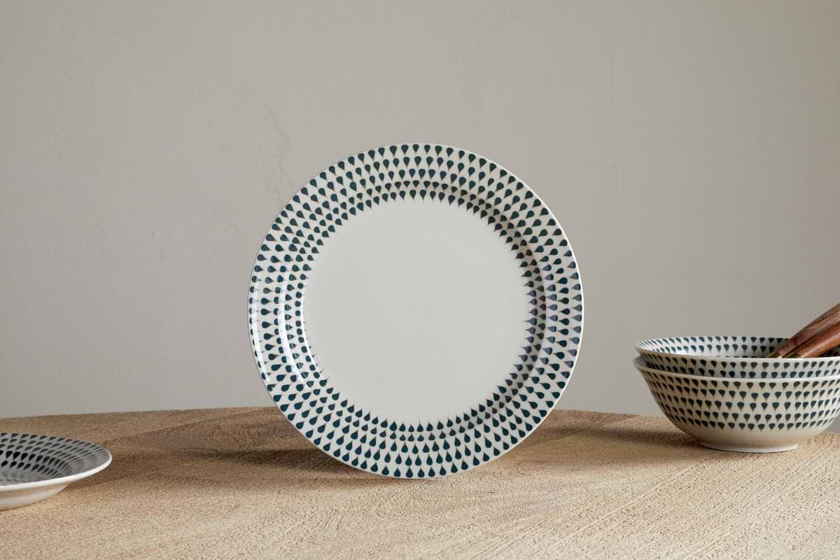 The nkuku Juniper Drop Side Plate in Cream & Juniper stands upright on a wooden surface, showcasing contemporary style. Matching bowls and a plate with a green drop pattern are partially visible nearby.