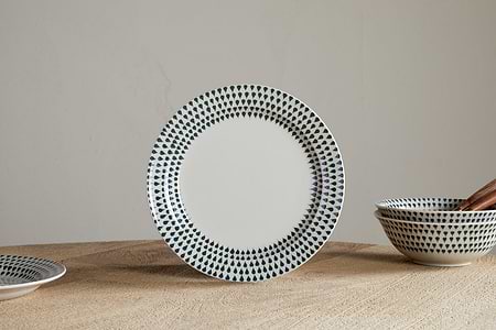 The nkuku Juniper Drop Side Plate in Cream & Juniper stands upright on a wooden surface, showcasing contemporary style. Matching bowls and a plate with a green drop pattern are partially visible nearby.