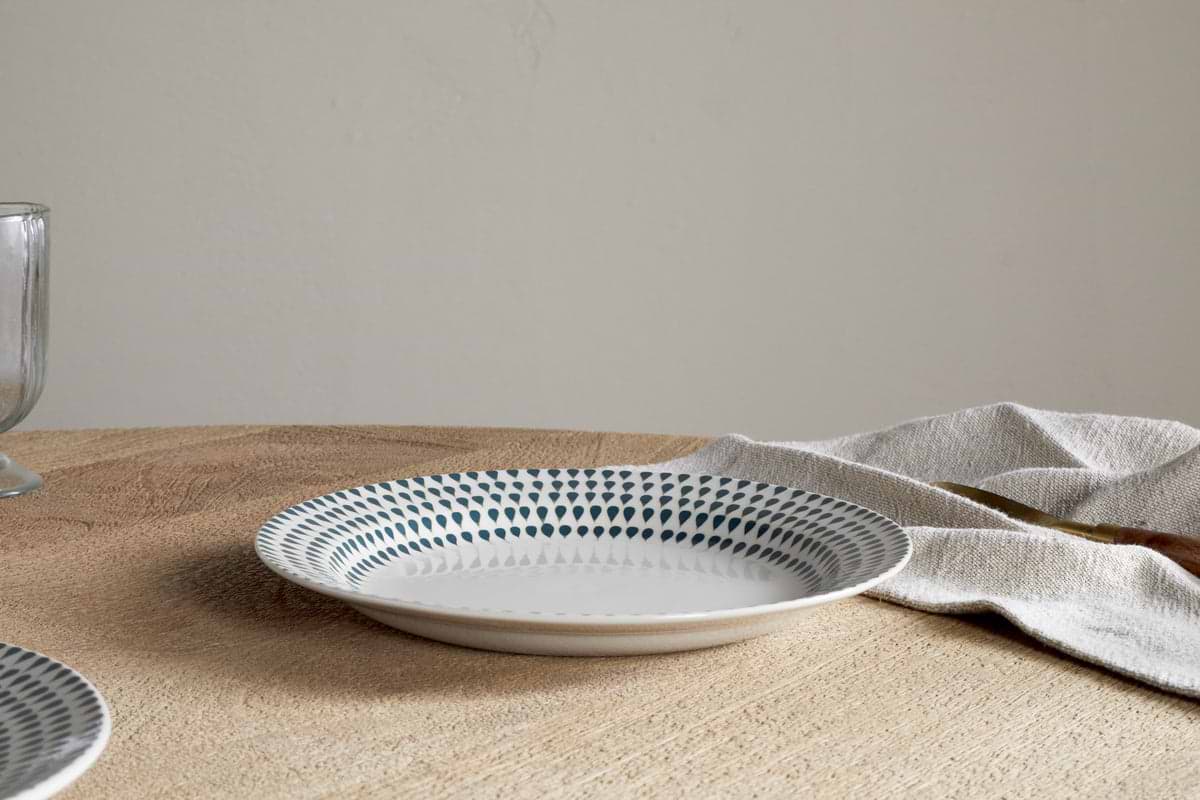 The nkuku Juniper Drop Dinnerware Set - Cream & Juniper (Set of 12) features a patterned ceramic plate with blue dots and subtle drop design on a wooden table beside a beige napkin; a glass appears partially on the left.