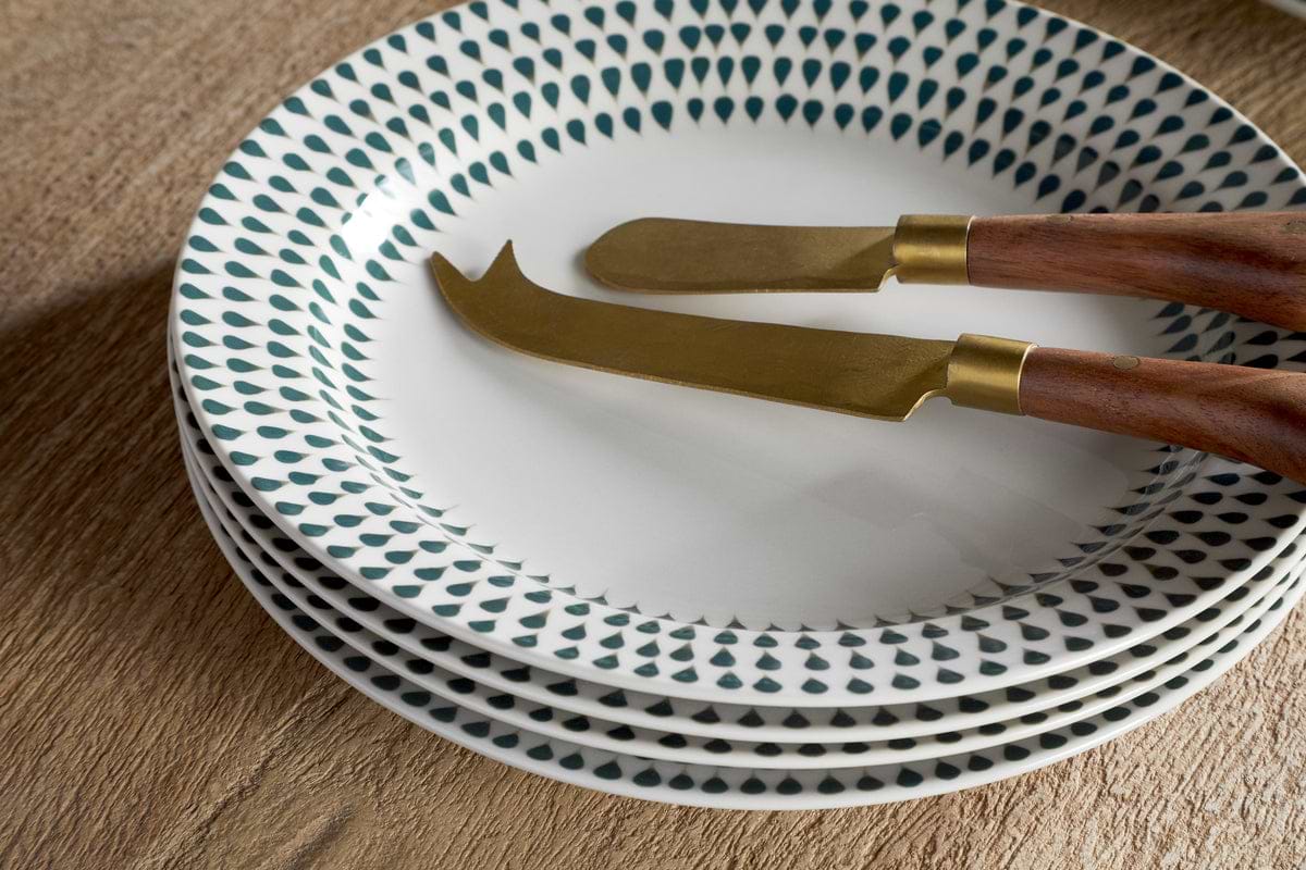 A stack of four nkuku Juniper Drop Side Plates in cream & juniper with a green drop rim rests on wood—ideal for modern dining. Two cheese knives with gold blades and wooden handles are placed on the top plate.