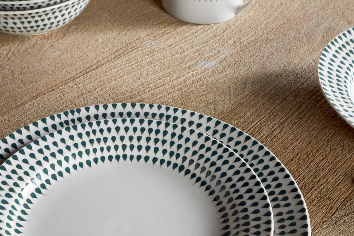Close-up of the nkuku Juniper Drop Dinnerware Set - Cream & Juniper (Set of 12), showing white ceramic plates and bowls with a juniper green design on wood; a matching mug and plate add to the elegant set in the background.