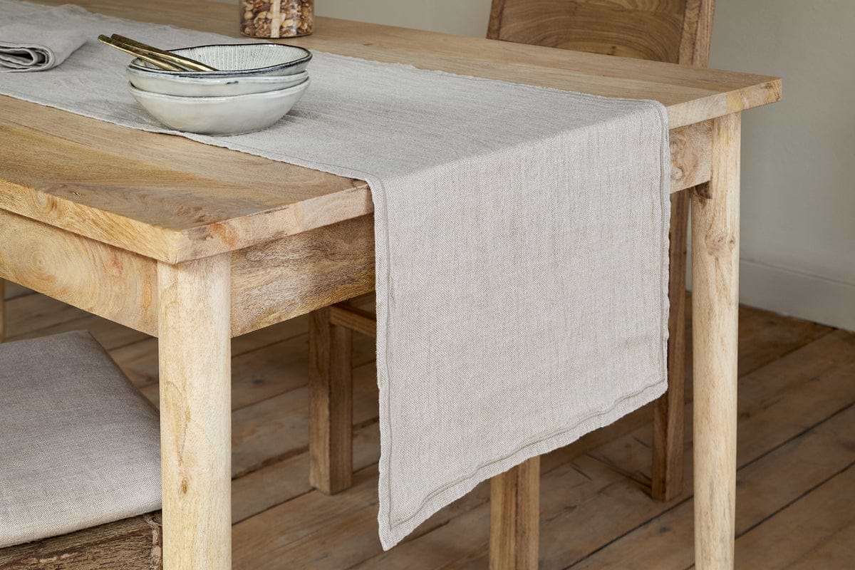 A cozy, minimalist room features a light wooden dining table topped with the nkuku Junto Linen Runner in Natural (Large), a matching bowl with utensils, and a wooden chair with a linen cushion on a wooden floor.