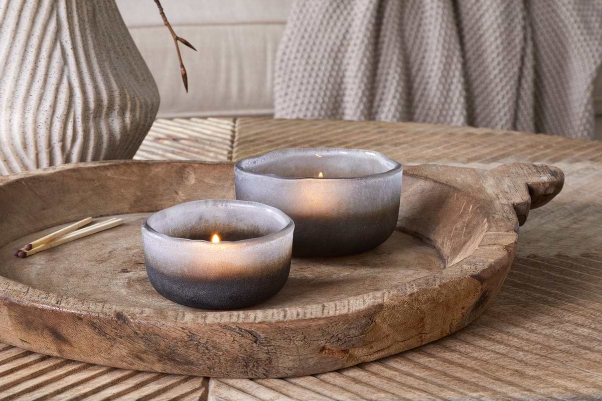 Two Janka Tealight Holders - Smoked Charcoal (Set of 2) by nkuku glow on a rustic wooden tray beside matches, a textured beige vase, and a knitted taupe blanket—perfect accents for cozy, neutral home decor.