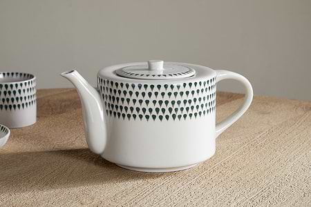 The nkuku Juniper Drop 4 Person Teapot in Cream & Juniper, featuring a round handle and spout and decorated with dark teardrop motifs, sits on a light wood surface—an elegant hand-painted gift for tea lovers, with a matching cup behind.