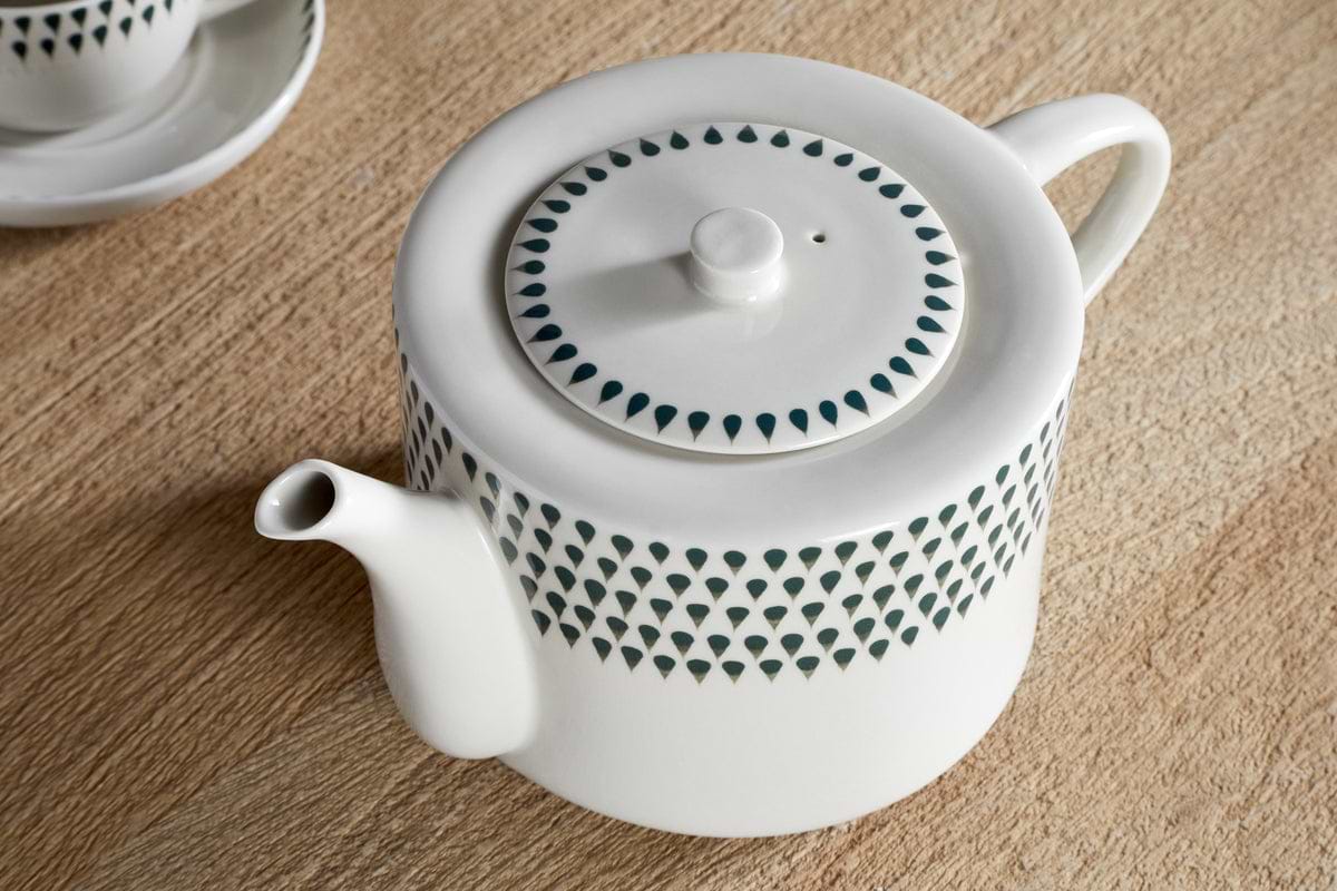 The nkuku Juniper Drop 4 Person Teapot in Cream & Juniper, hand-painted with dark green teardrop motifs, sits on a wooden surface. A matching saucer appears in the background—an ideal gift for tea lovers.