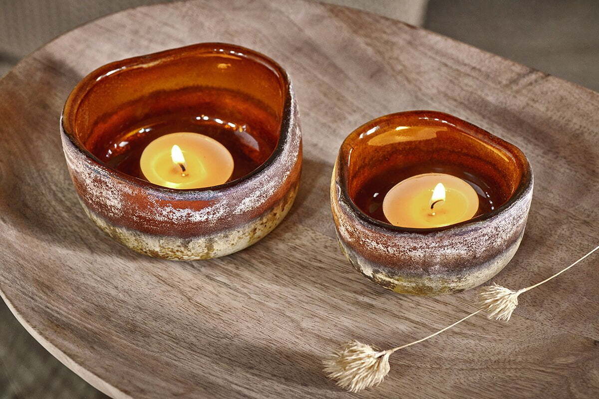 Two lit Janka Tealight Holders - Dark Amber by nkuku, crafted in rustic brown ceramic with uneven edges, sit atop a round wooden tray. A single dried grass stem nearby adds a natural accent to the cozy display.