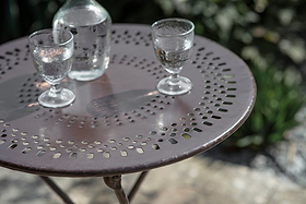 The Jeavika Iron Outdoor Bistro Table by nkuku in black holds two stemmed water glasses and a glass bottle. Sunlit among greenery and stones, it creates a relaxed outdoor vibe with its decorative metal design.