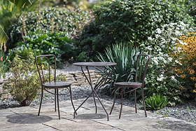 The nkuku Jeavika Iron Outdoor Bistro Table in black, paired with two matching chairs, sits on a stone patio amid lush green bushes and vibrant blooms, making it perfect for outdoor living.