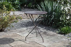 The Jeavika Iron Outdoor Bistro Table by nkuku stands on a stone patio amid greenery and white flowers. Its round black surface features a decorative pattern, and slender, collapsible legs make it perfect for stylish outdoor living.