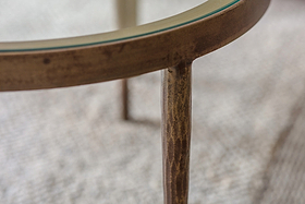 Close-up of nkuku's Jiya Iron & Glass Nested Coffee Tables - Brass, featuring an antique brass-finished metal frame and glass top. A carved leg is in focus on a textured rug.
