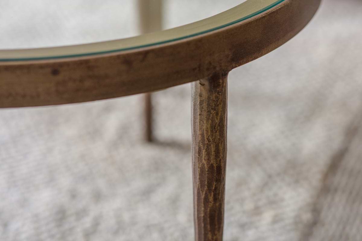 Close-up of nkuku's Jiya Iron & Glass Nested Coffee Tables - Brass, featuring an antique brass-finished metal frame and glass top. A carved leg is in focus on a textured rug.