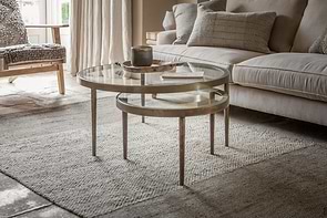 Two round nkuku Jiya Iron & Glass Nested Coffee Tables in brass sit on a woven beige rug in front of a light gray sofa with textured pillows. Soft natural light through sheer curtains creates a cozy, inviting vibe.