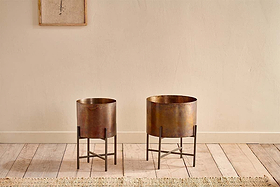 Two Juoni Iron Planters by nkuku, in bronze finish with black iron stands, are set side by side on a light wooden floor. Behind them is a cream wall with framed artwork and a woven rug at the base.