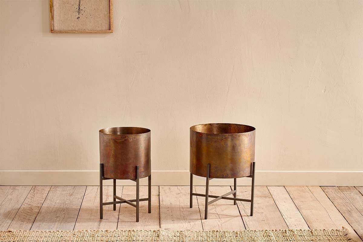 Two Juoni Iron Planters by nkuku, in bronze finish with black iron stands, are set side by side on a light wooden floor. Behind them is a cream wall with framed artwork and a woven rug at the base.