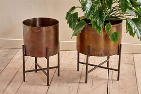 Two nkuku Juoni Iron Planters with a rustic bronze finish stand on dark iron stands atop a light wooden floor against a cream wall. One holds a lush green plant with large glossy leaves; the other is empty.