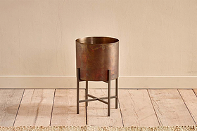 The Juoni Iron Planter by nkuku features a cylindrical, bronze-hued design on a black metal four-leg stand with crossbars, set on light wood flooring against a beige wall. A woven rug borders the scene—ideal for showcasing house plants.