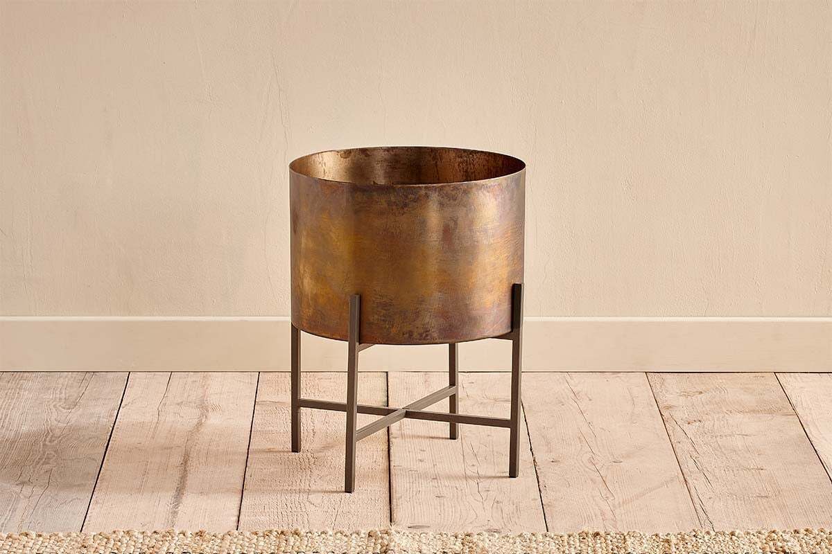 The Juoni Iron Planter by nkuku features a distressed brass finish and antique look, resting on a dark metal cross-shaped stand—perfect for house plants and beautifully suited for display on wooden floors.