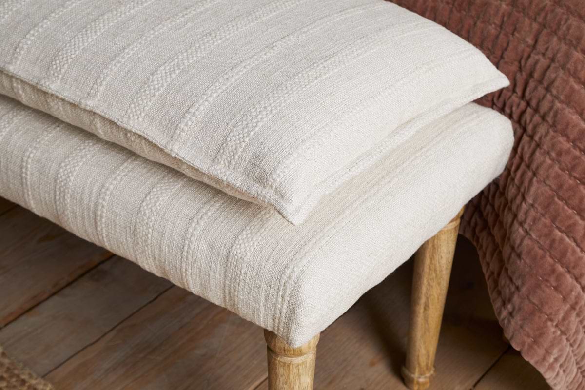 A close-up of the nkuku Kiana Cotton Upholstered Bench in Natural, topped with two cream textured cushions and set next to a rust-colored quilted blanket on a bed, all on a warm wooden floor.
