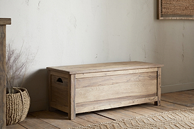 The Karadi Reclaimed Wood Storage Bench - Natural by nkuku rests on a wooden floor beside a woven basket and textured rug, all set against a light-colored wall.