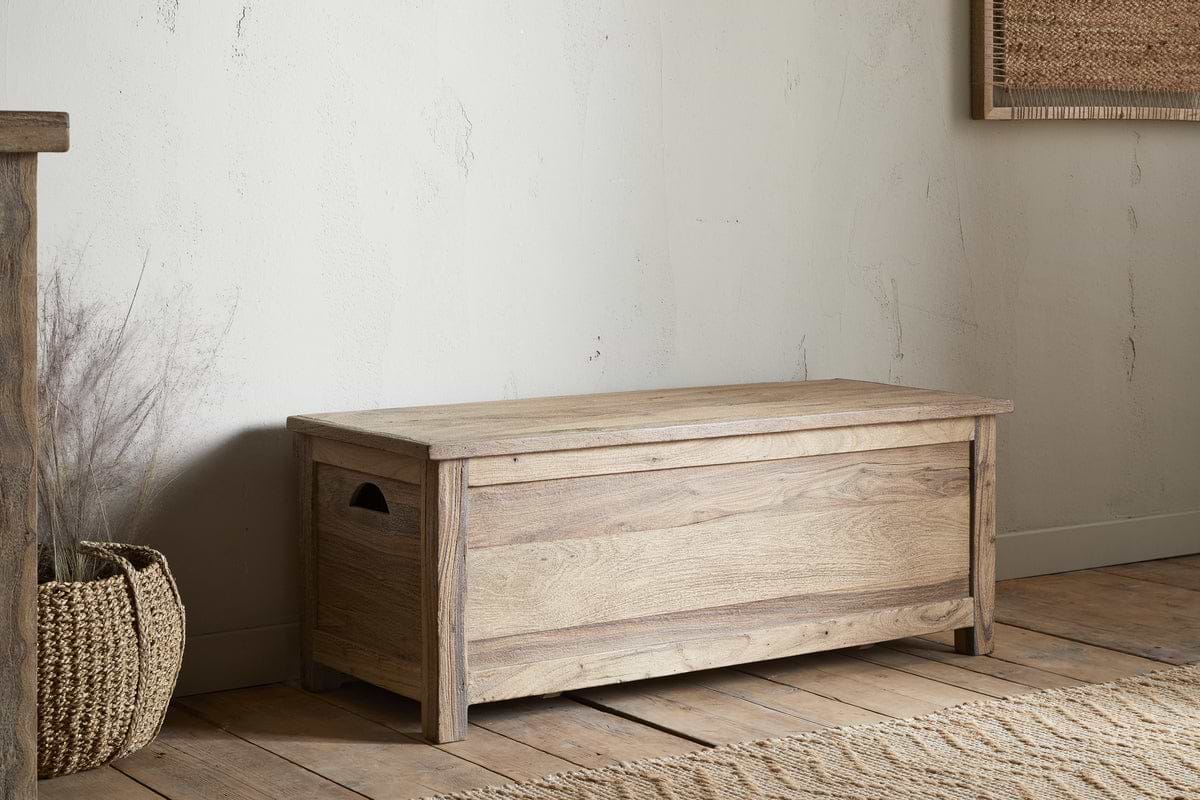 The Karadi Reclaimed Wood Storage Bench - Natural by nkuku rests on a wooden floor beside a woven basket and textured rug, all set against a light-colored wall.