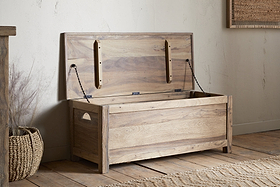 The Karadi Reclaimed Wood Storage Bench - Natural by nkuku sits open in a rustic room with wooden floors, a woven basket nearby, and neutral-colored walls.