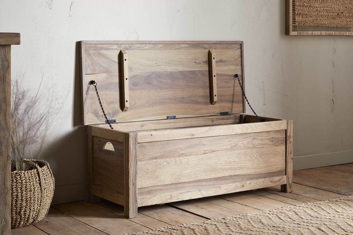 The Karadi Reclaimed Wood Storage Bench - Natural by nkuku sits open in a rustic room with wooden floors, a woven basket nearby, and neutral-colored walls.