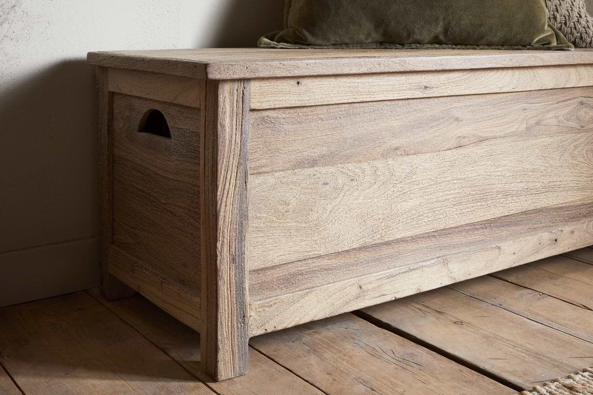 The Karadi Reclaimed Wood Storage Bench by nkuku features a hinged lid, cut-out handle for easy access, and a natural finish. It sits near a white wall on a wooden floor and is topped with a green cushion.