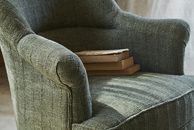 A close-up of the nkuku Kiana Cotton Armchair in Moss, featuring textured cotton upholstery, with two closed books stacked on one arm and bathed in soft natural light.
