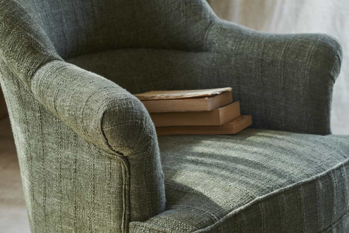 A close-up of the nkuku Kiana Cotton Armchair in Moss, featuring textured cotton upholstery, with two closed books stacked on one arm and bathed in soft natural light.