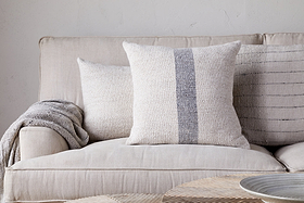 A beige sofa features the nkuku Kallu Wool & Cotton Cushion Cover in natural, a textured cream cushion, and a gray throw blanket. A light background wall and woven tray on the table finish this serene setting.