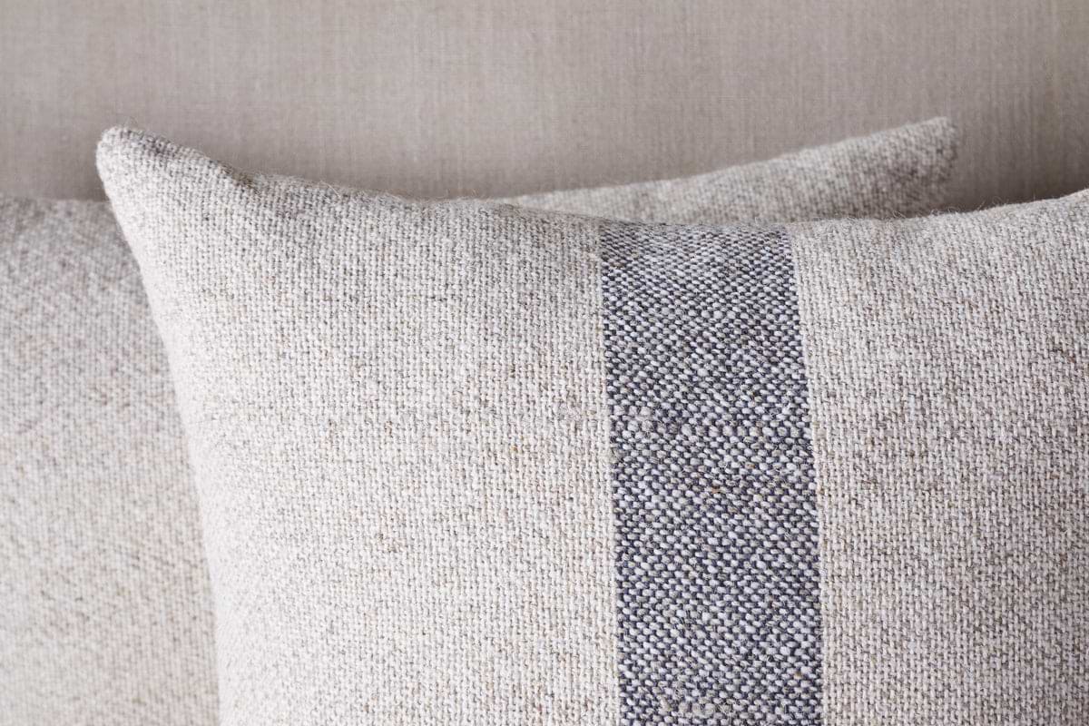 Two Kallu Wool & Cotton Cushion Covers by nkuku in natural tones, each with a central vertical blue-grey stripe, are arranged one behind the other against a matching fabric background.