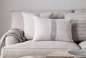 A beige sofa with nkuku’s Kallu Wool & Cotton Cushion Cover - Natural and three neutral throw pillows sits against a light wall, a gray knit blanket over the armrest. A round, light wood table appears in the foreground.