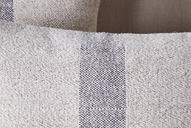 Close-up of the nkuku Kallu Wool & Cotton Cushion Cover - Natural, showing its textured beige fabric with a central vertical dark gray and white stripe. A similar cushion is softly blurred in the background.