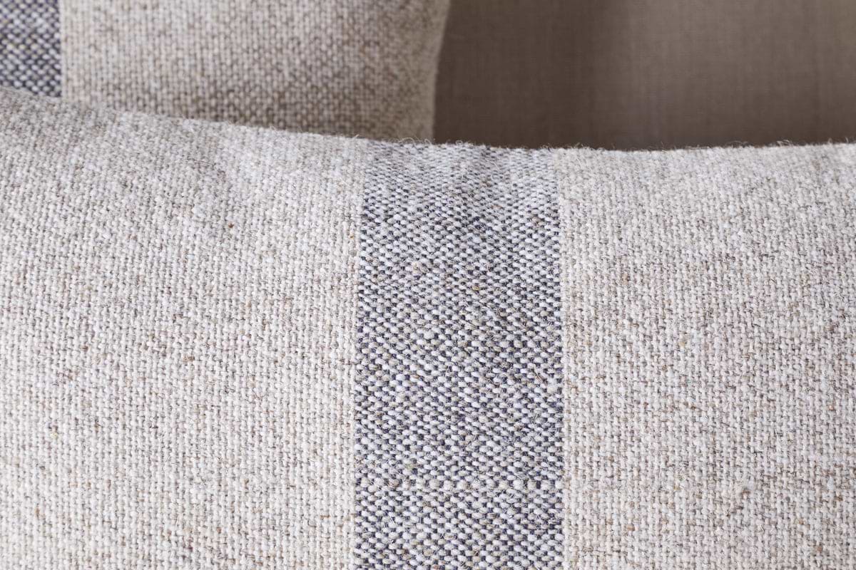 Close-up of the nkuku Kallu Wool & Cotton Cushion Cover - Natural, showing its textured beige fabric with a central vertical dark gray and white stripe. A similar cushion is softly blurred in the background.