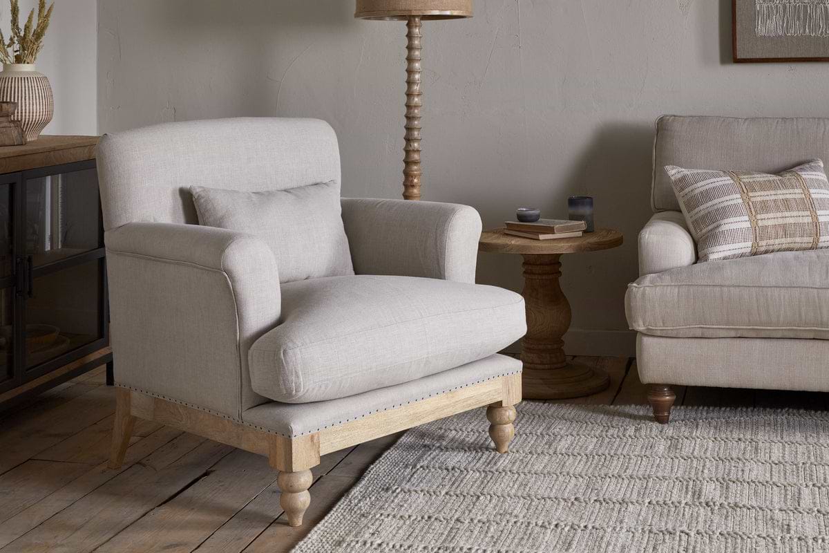A neutral-toned living room features the nkuku Kashi Linen Armchair in natural with wooden legs, a matching pillow, a round solid mango wood side table, a beige sofa, and a textured rug on wooden flooring.