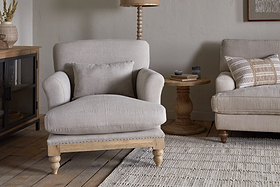 A cozy living room featuring the Kashi Linen Armchair - Natural by nkuku with a matching pillow, a round solid mango wood side table, part of a beige sofa, a textured rug, and wood flooring in neutral decor.